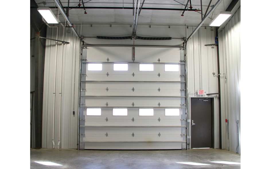 Interior view of an industrial garage with a large Thermacore Model 850 roll-up door featuring six small windows, metal walls, a gray door with an exit sign, and bright overhead lighting.