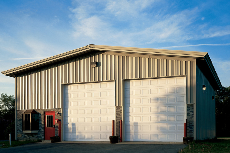 A metal building with two large white Sectional Steel Doors (Model 594), a small red entrance door, a window, and two planters in front. The building sits under a blue sky with some clouds, lit by evening sunlight.