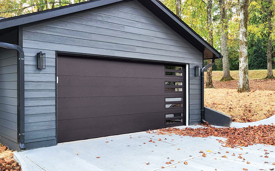 A modern gray garage with a dark brown Overhead Door from the Thermacore Collection, featuring horizontal glass panels on the side, surrounded by trees with fallen autumn leaves on the concrete driveway.