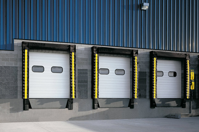 Three white Thermacore Sectional Steel Doors, Model 591, with yellow and black bumpers are set in a gray and blue industrial building wall, each featuring small rectangular windows.
