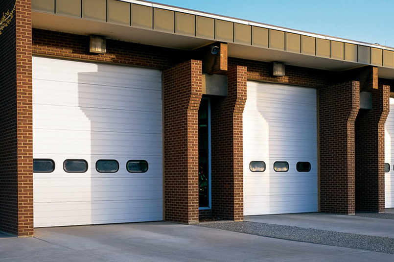 Two large white Thermacore sectional doors with small horizontal windows, set in a brick building with concrete pavement in front, under a blue sky.