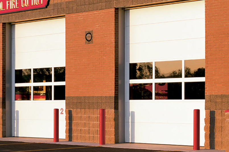 Two large white Thermacore sectional steel doors on a brick building, likely a fire station, with red bollards in front and sunlight reflecting in the windows. The number 2 is visible near one of the doors.