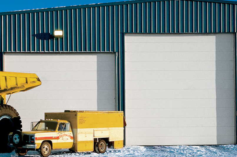 A yellow utility truck and the wheel of a large construction vehicle are parked outside a blue metal building with two large white Sectional Steel Doors on a snowy ground.