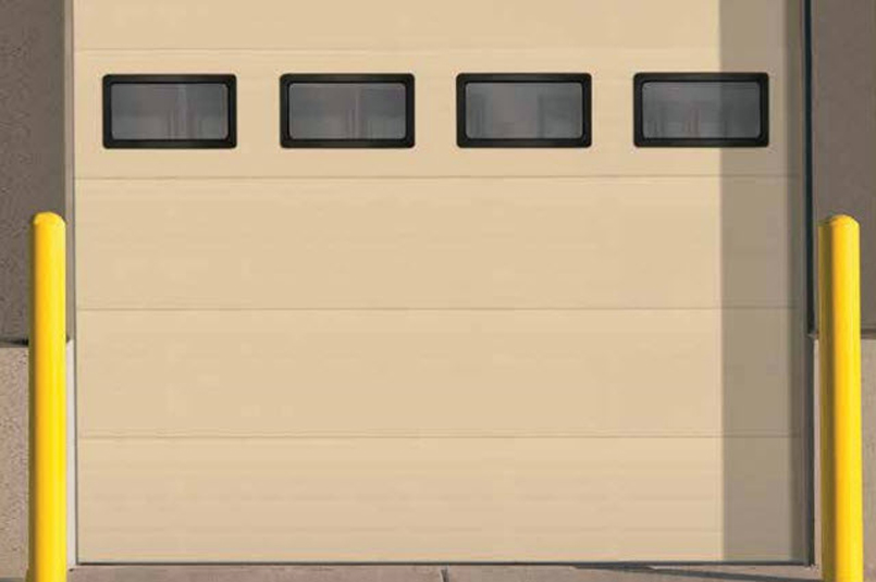 A closed beige Thermacore industrial garage door, Model 850, features three rectangular windows near the top and two yellow protective bollards on either side of the entrance.