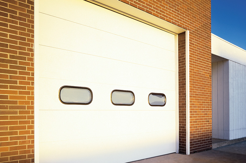 A large white Model 598 sectional steel door with three small rectangular windows is set in a brick building. The structure is partially visible, with a clear blue sky in the background.