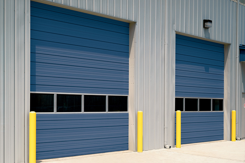 Two large blue Sectional Steel Doors with horizontal panels and small windows near the bottom, set in a light gray metal building. Yellow safety bollards stand in front of each door on a concrete surface. Model 432 shown.