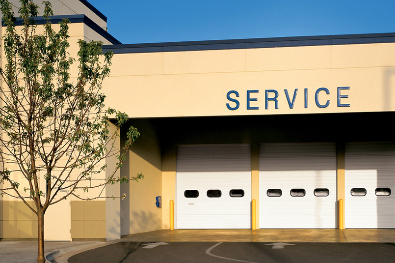 A beige building with the word SERVICE above two closed Model 598 sectional steel doors, a tree to the left, and yellow bollards in front of the doors on a sunny day.