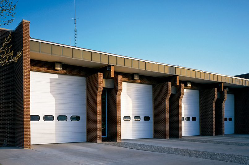 A row of four large, white Thermacore sectional steel doors—likely Model 598—on a brick building, possibly a fire station or service facility, stands out on a clear day with blue sky.