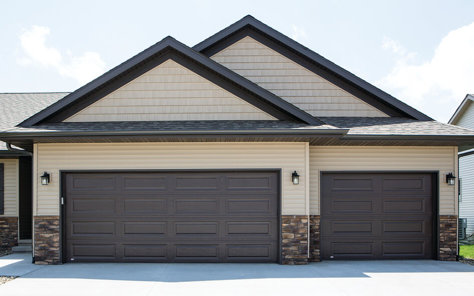 A modern house with tan siding, stone accents, and a concrete driveway features steel garage doors in dark brown, along with two prominent gable roofs.