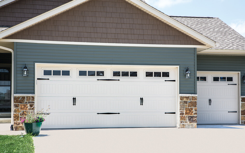 A house exterior with two large steel garage doors featuring decorative black hardware and windows at the top. The house has blue siding, brown shingles, stone accents, and two wall lanterns. A green potted plant sits nearby.