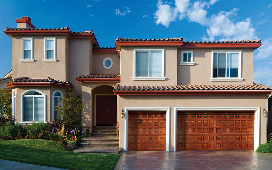 Two-story beige stucco house with red tile roof, large windows, traditional wood double front doors, and a three-car garage with wooden doors. Well-maintained lawn and landscaping out front under a blue sky with scattered clouds.