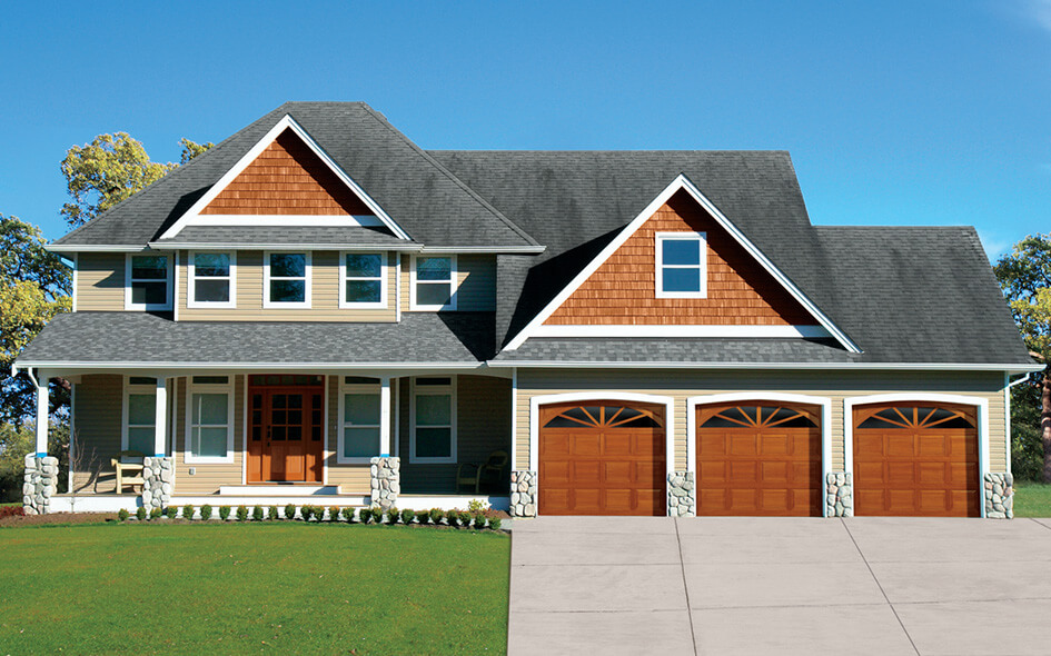 A two-story suburban house with beige siding, white trim, and dark gray roof. The front features a porch with columns and three traditional wood garage doors. The house is surrounded by green lawn and trees under a blue sky.