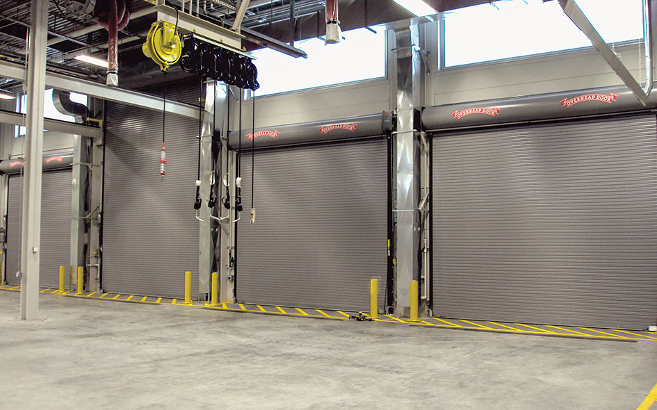Three large industrial roll-up doors inside a warehouse, equipped with RSX Fire Door Operators. Yellow bollards and striped caution lines are in front, with overhead equipment and pipes visible and natural light streaming from high windows.