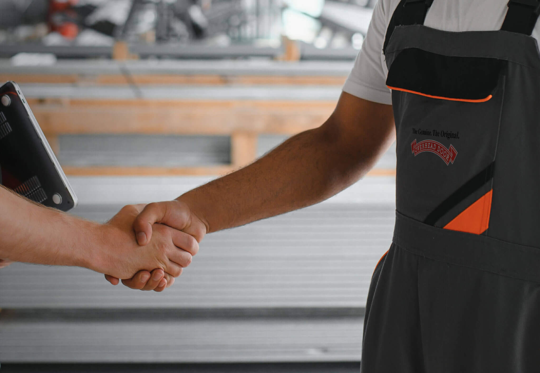 Two people shake hands—one in a dark work uniform with orange accents and text, the other holding a digital tablet—suggesting a discussion about Maintenance Programs. The background features blurred industrial or construction materials.