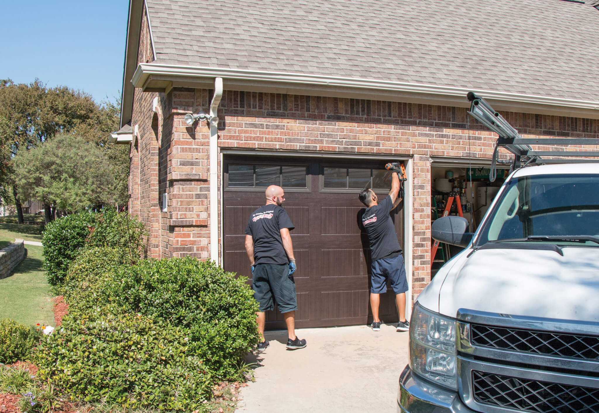 Two men in black shirts work on a closed brown garage door of a brick house, tools nearby and a white truck parked in the driveway. With shrubs and trees surrounding the area, they appear to be performing garage door Maintenance Programs.