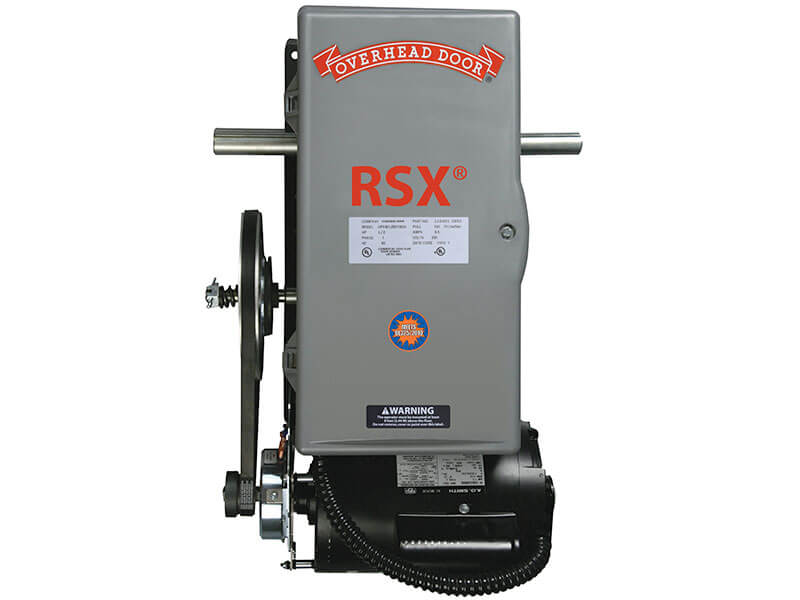 Gray industrial garage door opener labeled RSX with the Overhead Door logo at the top. The device has various warning labels, exposed wiring, and mechanical components on the left side.