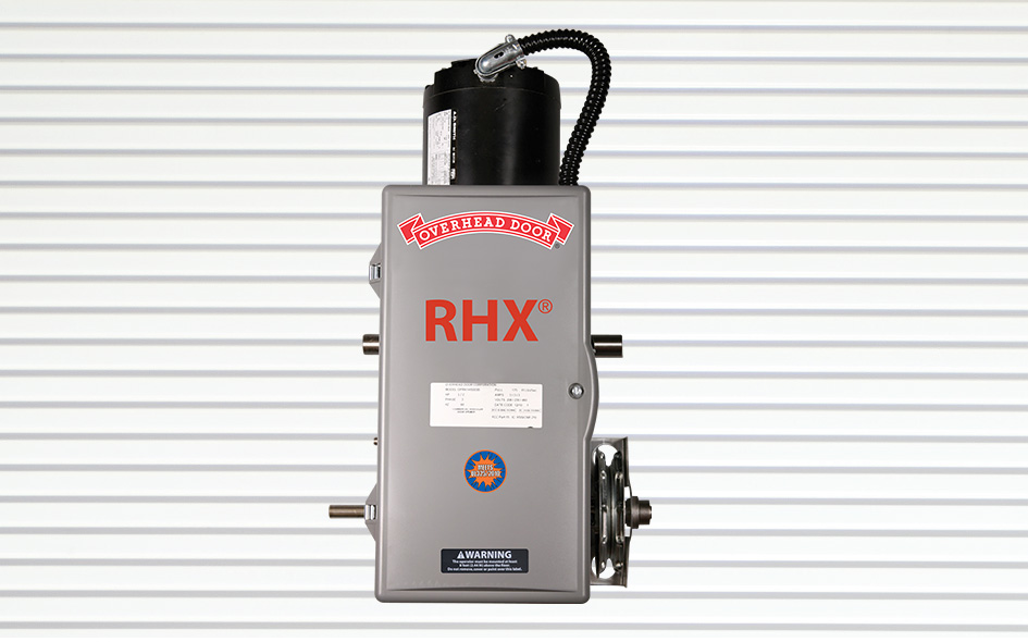 Gray RHX door operator for commercial garage door opener applications, mounted on a white, horizontally-striped background. The device features various labels, a red logo, and a black motor on top.