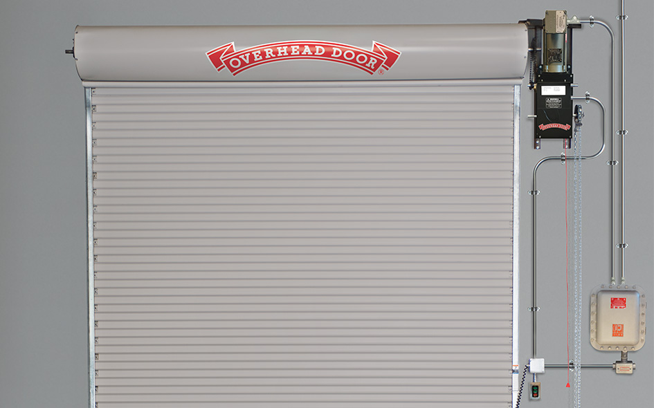 A gray metal roll-up overhead door with the words Overhead Door in red on top, installed in a building with a visible RHX Explosion-Proof Operator and control box mounted on the right side of the door.