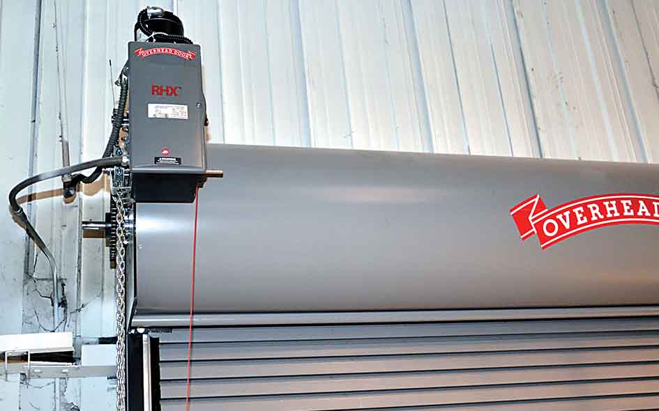 A close-up of a gray commercial roll-up garage door with an RHX Commercial Door Operator mounted to the wall. The operator features a chain mechanism and is labeled Overhead Door and RHX. The door is partially closed.