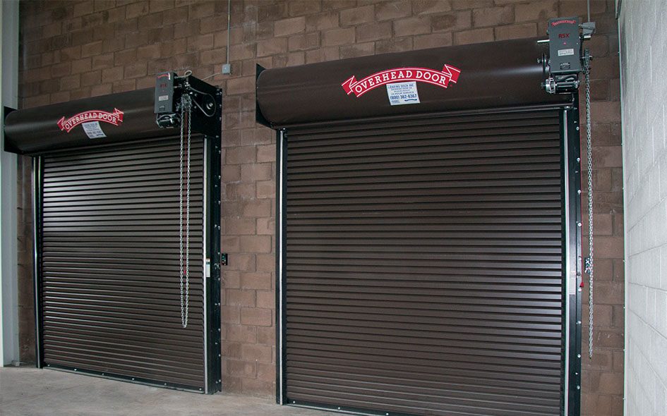 Two brown metal rolling overhead doors with red Overhead Door logo signs are installed on a brick wall inside the building, each equipped with a Commercial Door Operator RSX mounted above for smooth, reliable operation.