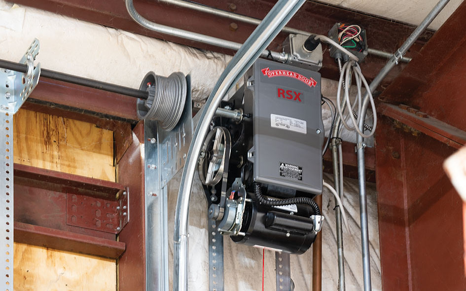 A Commercial Door Operator labeled RSX is mounted on a metal frame, surrounded by various wires, metal conduits, and a pulley system in an unfinished building.