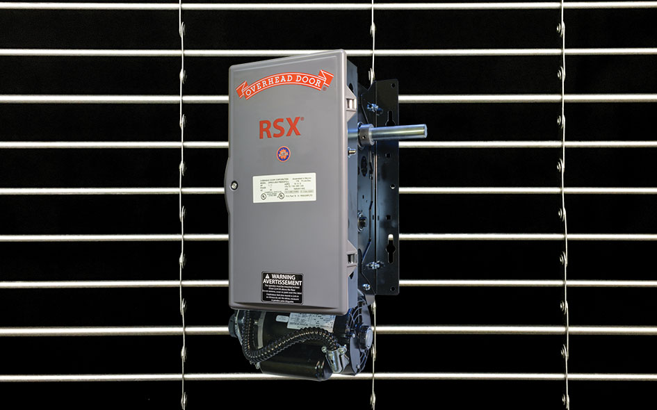 A gray RSX Egress Operator garage door unit is mounted against a black backdrop with horizontal white lines. The operator features a label and warning stickers, with exposed wiring visible at the bottom.