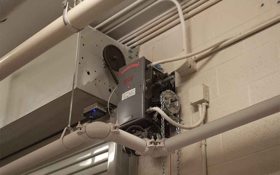 A Commercial Door Operator RSX with visible gears, chains, and electrical wiring is mounted on a white brick wall near ceiling pipes.