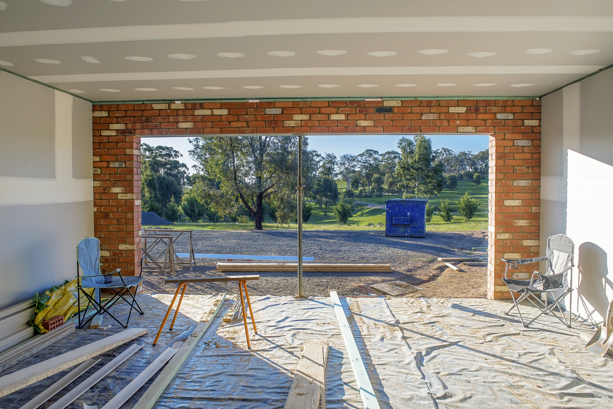 A partially constructed room with exposed brick and unfinished walls opens to a sunny outdoor area with trees, a blue dumpster, and construction materials scattered on the ground. Two folding chairs and a stool sit inside, next to where a fire rated garage door may be installed.