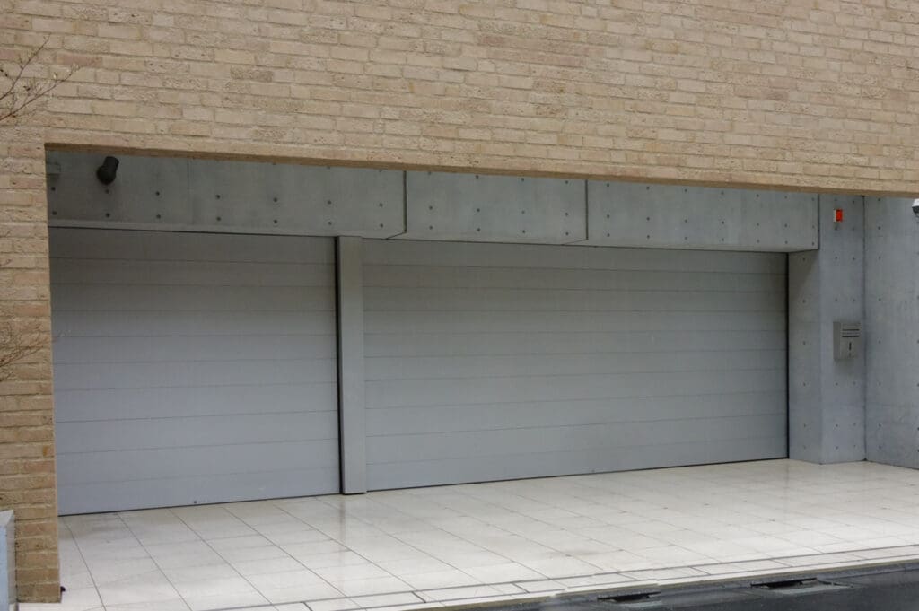 A modern double garage features two closed, light gray fire rated garage doors set in a beige brick and concrete building. The floor in front is tiled in white, and there are no cars or people present, ensuring both style and fire safety.