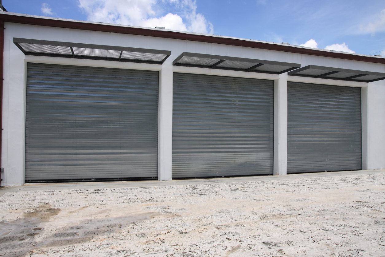 Three large closed metal roller shutter doors, each meeting fire rated garage door requirements, are set on a white industrial building with a concrete ground below and blue sky with scattered clouds above.