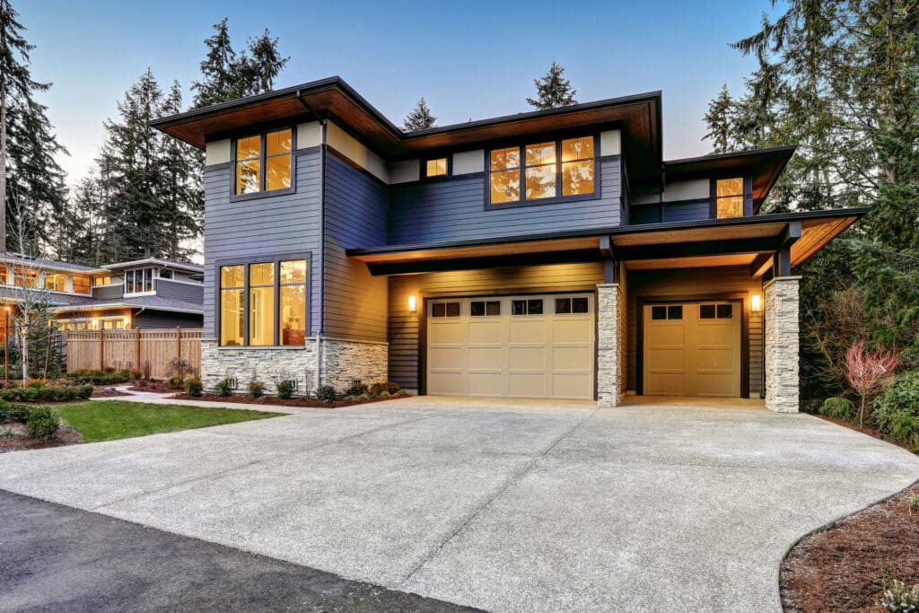 Modern two-story home with large windows, dark blue siding, stone accents, and a double garage. The spacious driveway highlights the home's curb appeal—perfect for those seeking seamless garage door repair or replacement at dusk amid lush landscaping.