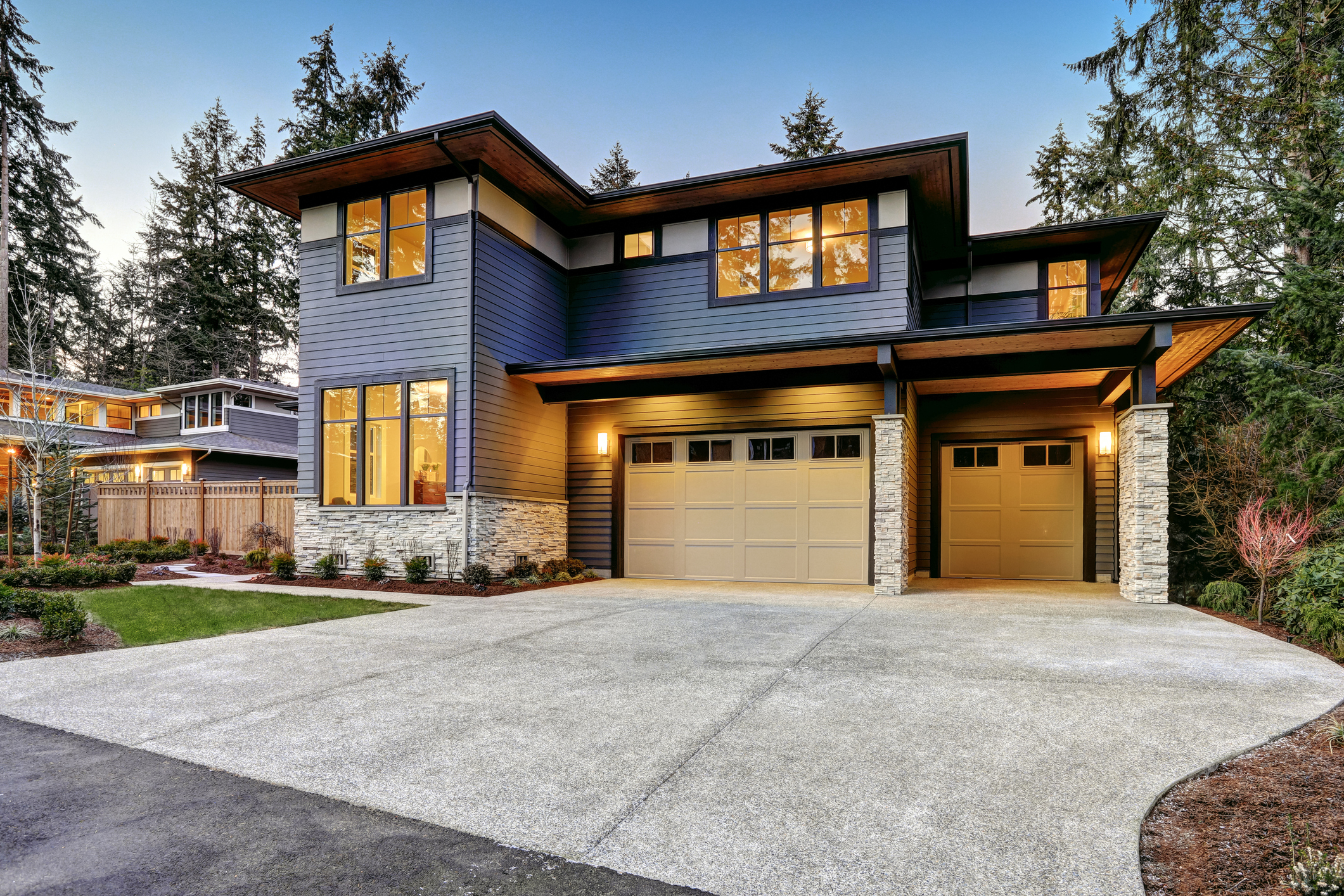 Modern two-story home with large windows, dark blue siding, stone accents, and a double garage. The spacious driveway highlights the home's curb appeal—perfect for those seeking seamless garage door repair or replacement at dusk amid lush landscaping.