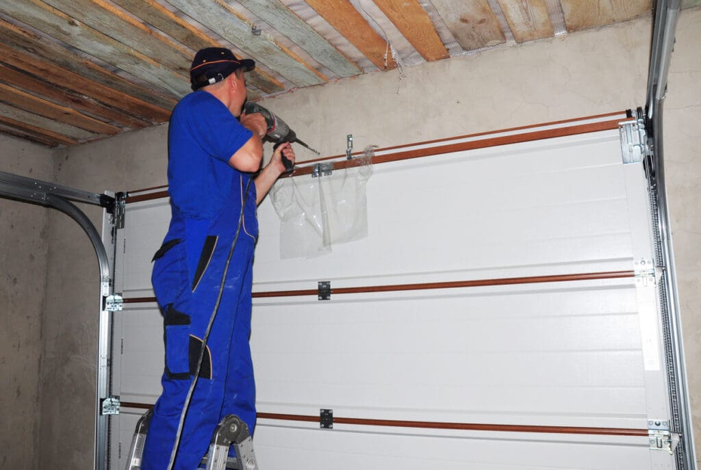 A person in blue overalls stands on a ladder, using a power drill to handle garage door repair vs replacement on a white garage door inside a partially finished room.