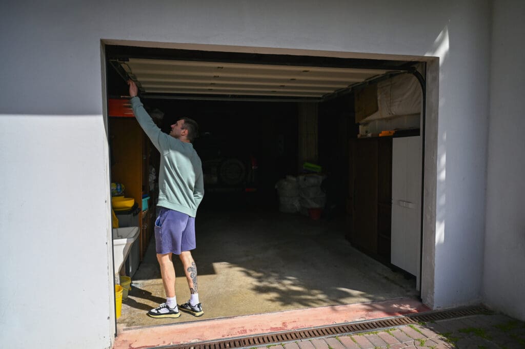 A person in a long-sleeve shirt and shorts stands at the entrance of an open, fire rated garage door, reaching up to pull it down. Sunlight casts shadows on the white exterior wall.