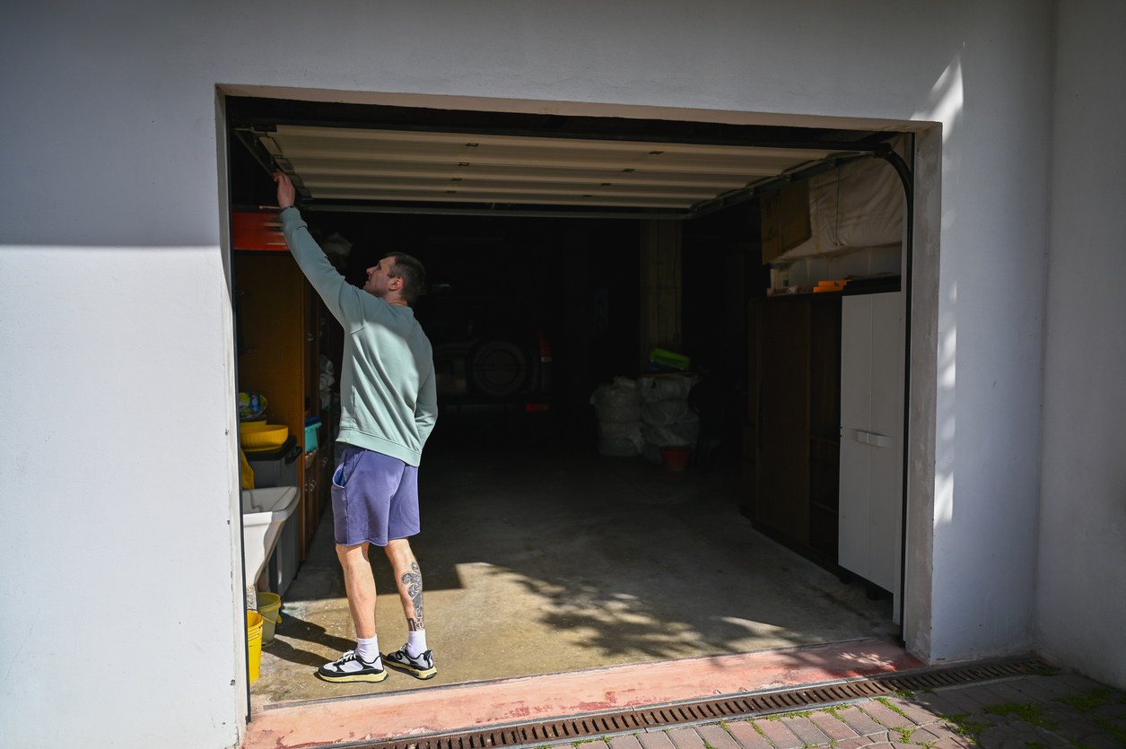 A person in a long-sleeve shirt and shorts stands at the entrance of an open, fire rated garage door, reaching up to pull it down. Sunlight casts shadows on the white exterior wall.