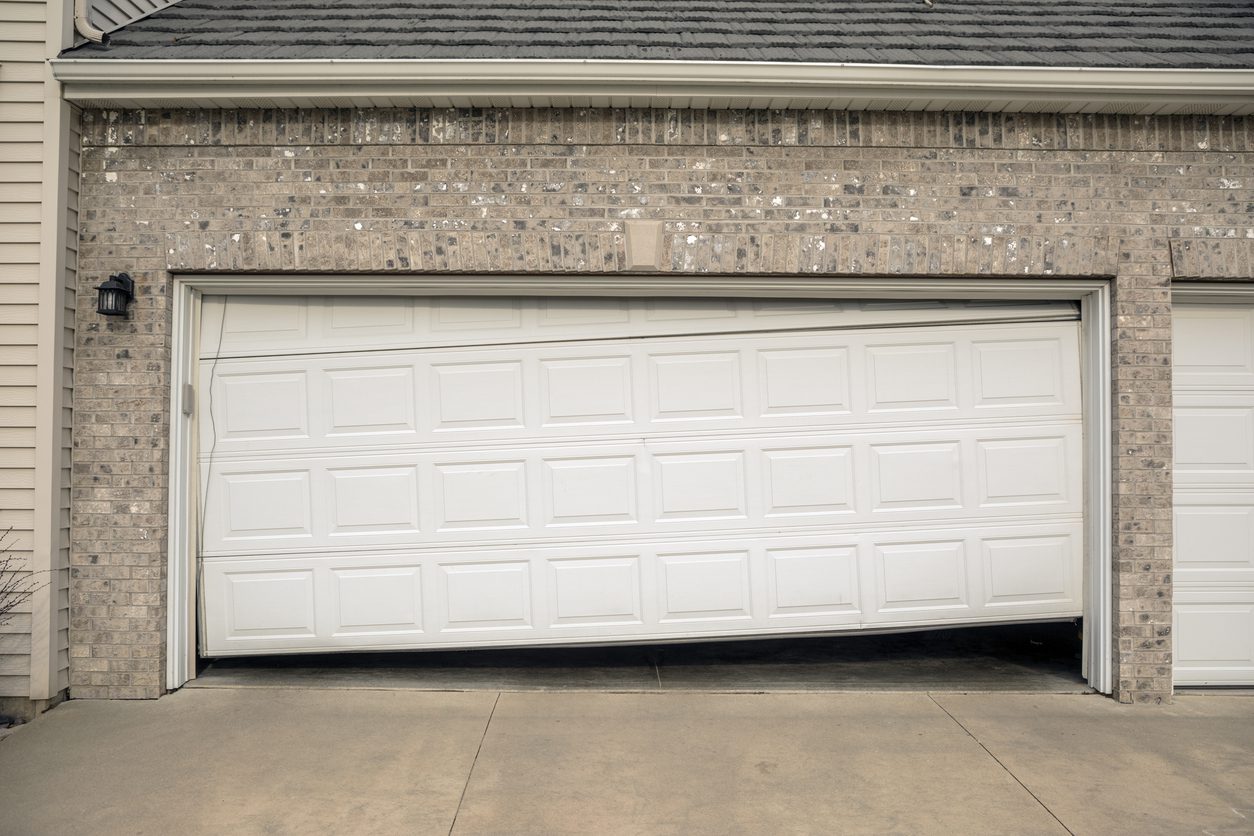 A white garage door is stuck halfway open and appears crooked, with one side higher than the other, on a brick house—highlighting the need to consider garage door repair vs replacement. The driveway and part of the garage interior are visible.
