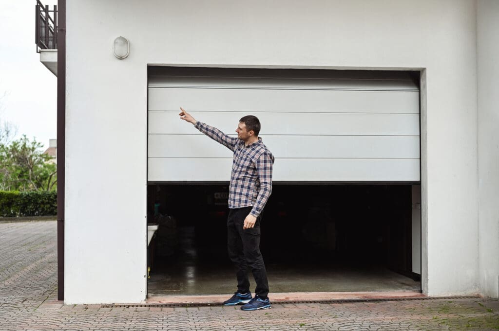 A man in a plaid shirt and black pants stands in front of a partially open garage door, pointing upward toward a light fixture—perhaps considering garage door repair vs replacement options.