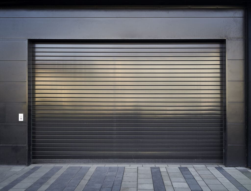 A closed, dark gray metal garage door with horizontal lines, set in a modern black wall. The design may meet fire rated garage door code, while alternating light and dark rectangular tiles line the ground. A control button is mounted on the left side of the door.